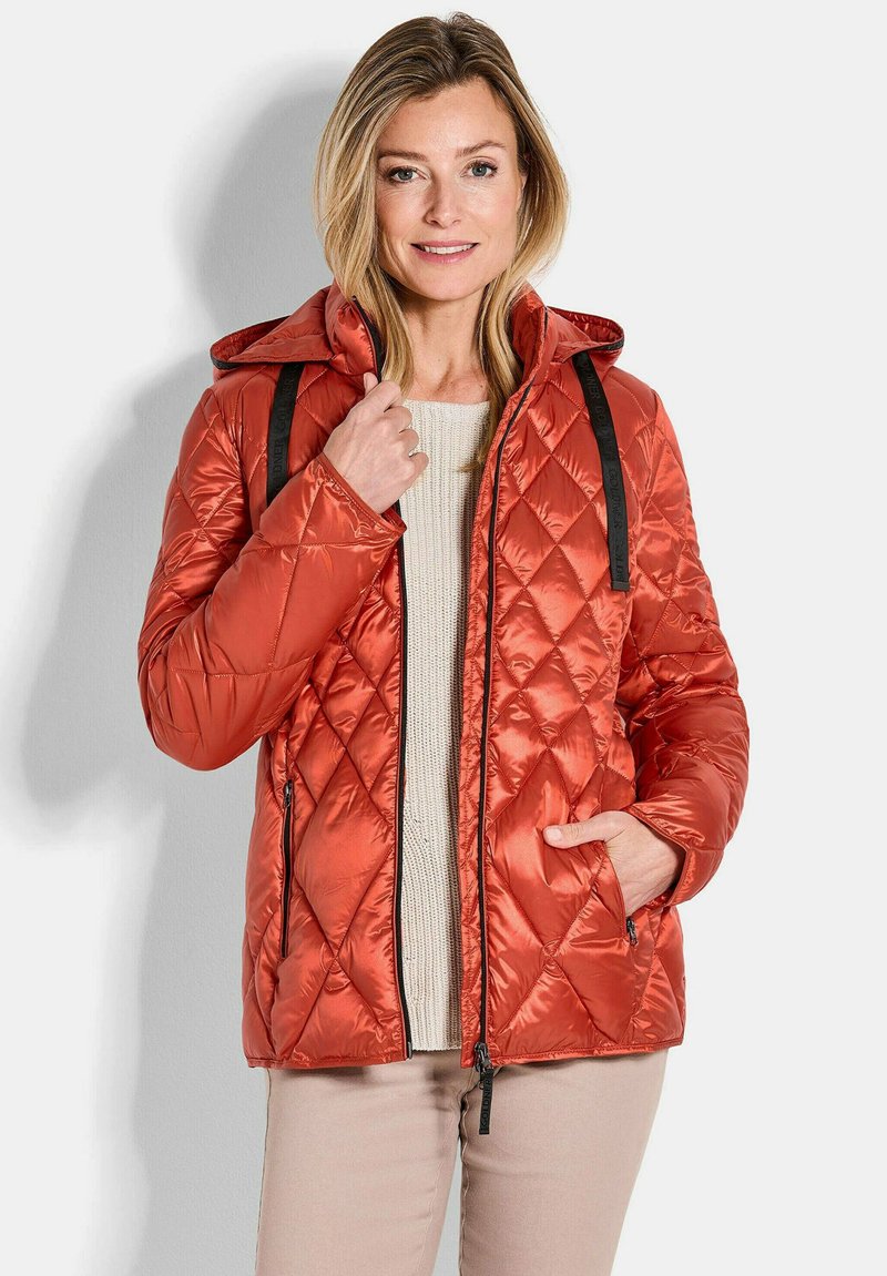 Gequilted, gewatteerde oranje jas met een capuchon; heeft zwarte ritsen en banden, en een gestructureerde, glanzende afwerking. Draagt over een crèmekleurige trui.