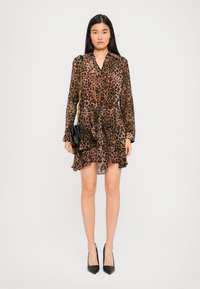 Kvinna klädd i en klänning med brun och svart leopardmönster med långa ärmar, håller en svart handväska med guldkedja, och bär svarta klackar.