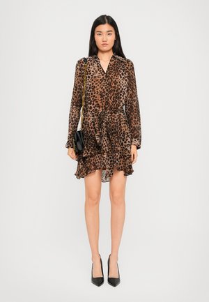 Femme portant une robe léopard marron et noir avec des manches longues, tenant un sac à main noir avec une bandoulière en chaîne dorée, et portant des talons noirs.