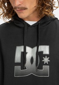 A man with curly hair and a mustache wears a black hoodie featuring a large silver logo, with two drawstrings visible near the neckline.