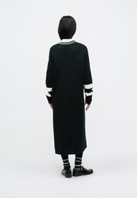 Dolga črna pletena obleka z belim ovratnikom in rokavi s črtami, ohlapen kroj, sega do sredine meč. Oblečena s črtastimi nogavicami in črnimi čevlji.