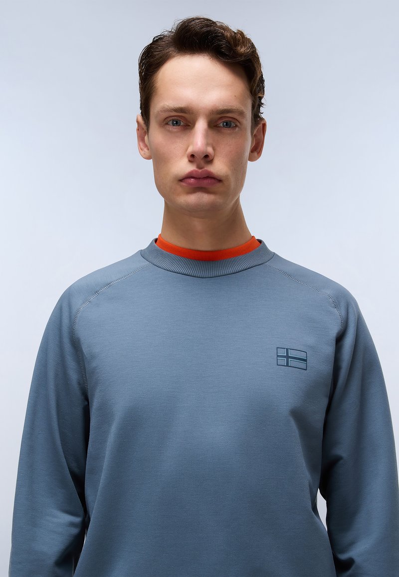 Grauer Langarmpullover mit Rundhalsausschnitt und Raglanärmel, mit einem orangen Innenkragen und einem kleinen Logo auf der Brust.