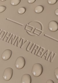 Tappeto beige texturizzato con gocce sollevate e logo impresso "JOHNNY URBAN", con un elemento di design circolare e finitura liscia.