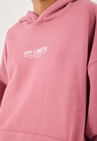 Person med en rosa hettegenser med hvit tekst "OFF LIMITS" og mindre tekst "DANGEROUS AREA" på brystet.