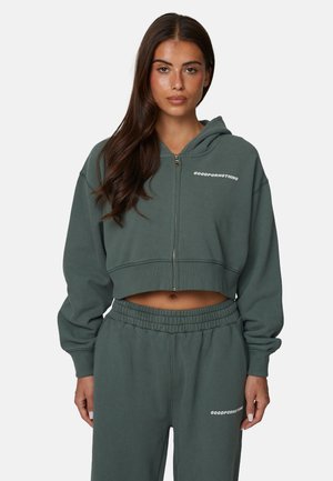 Kobieta o długich brązowych włosach, ubrana w ciemnozieloną, krótką bluzę na suwak i pasujące spodnie dresowe z białym napisem "GOODFORNOTHING".