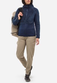 Kobieta w granatowej polarowej kurtce i khaki spodni cargo, nosząca dużą beżową torbę na jednym ramieniu, stojąca z nogami skrzyżowanymi.