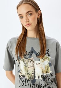 Camiseta gris de manga corta con un diseño gráfico de cuatro gatos y texto que dice: "¿A dónde miran estos gatos?" en un tipo de letra en negrita.