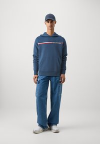Sudadera navy con letras de rayas rojas y blancas, combinada con pantalones cargo de denim de pierna ancha. El modelo lleva una gorra y zapatillas a juego en navy.