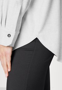 Nahaufnahme einer linken Hand, die an einer schwarzen Hose ruht. Die Person trägt ein hellgraues Hemd mit geknöpften Manschetten und einem abgerundeten Saum.