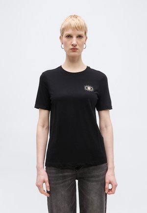 Γυναίκα με κοντά ξανθά μαλλιά φοράει μαύρο μπλουζάκι με λευκό γραφικό μάτι στο στήθος και ξεβαμμένο μαύρο τζιν, στέκεται μπροστά σε απλό ανοιχτό φόντο.