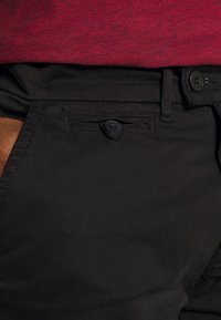 Gros plan sur un pantalon noir avec une petite poche boutonnée et des passants de ceinture, porté avec une chemise rouge texturée rentrée.