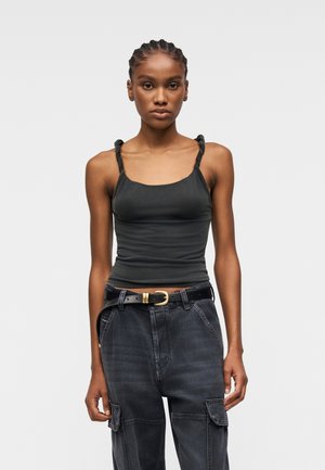 Fonott hajú nő szűk, fekete, csavart pántos atlétatrikóban és sötét cargo farmert visel, fekete övvel.