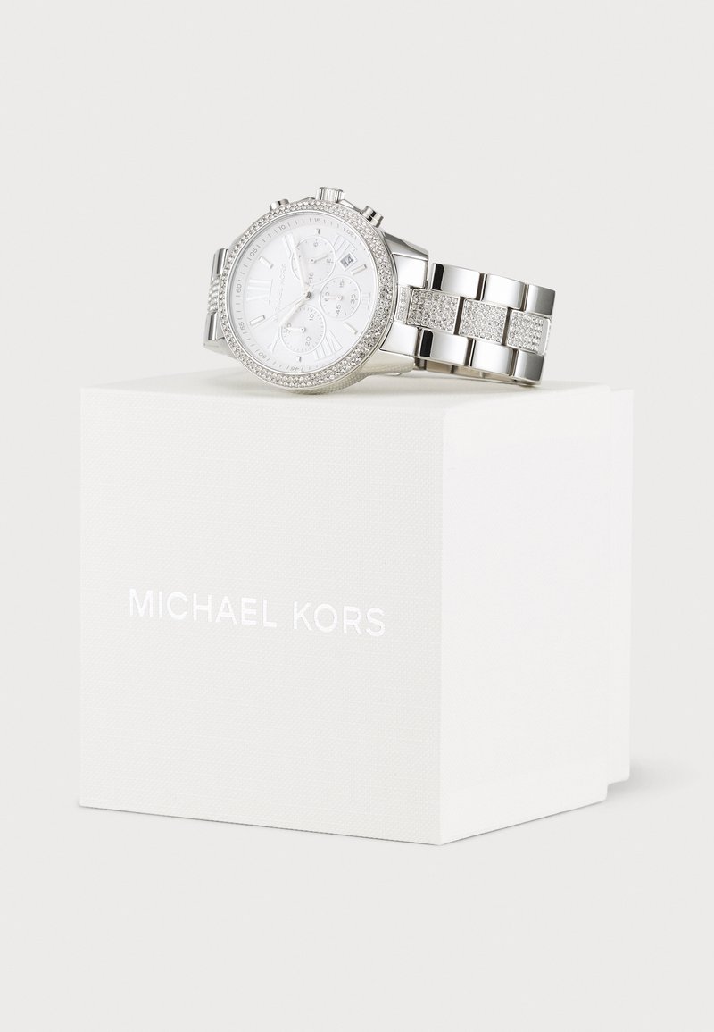 Montre chronographe argentée avec un cadran blanc, des accents en pierre et un bracelet en métal, présentée sur une boîte blanche avec le texte "MICHAEL KORS".