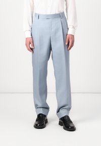 Pantalones plisados de color azul claro, fabricados en una tela suave, con un corte entallado y una abertura de pierna amplia, combinados con zapatos negros.