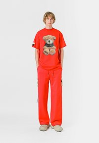 Camiseta roja con un gráfico de un oso de peluche, combinada con pantalones rojos a juego. El conjunto cuenta con dos bolsillos laterales con acentos negros y calzado texturizado.