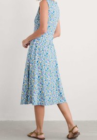 Kobieta w bezrękawowej sukience do kolan w niebiesko-fioletowo-żółty kwiatowy wzór, nosząca brązowe płaskie sandały, stojąca na szarym podłożu.