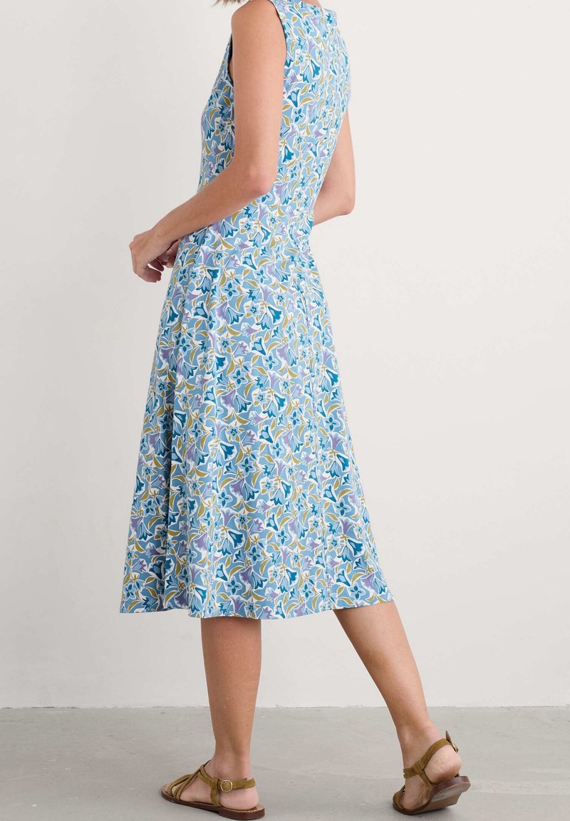 Kobieta w bezrękawowej sukience do kolan w niebiesko-fioletowo-żółty kwiatowy wzór, nosząca brązowe płaskie sandały, stojąca na szarym podłożu.