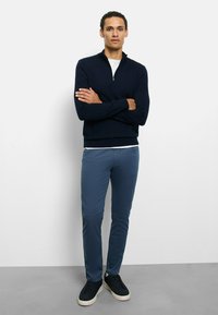 Marineblauer Reißverschluss-Pullover, hellgraues Hemd darunter, blaue Slim-Fit-Hose und schwarze Sneaker mit weißen Sohlen. Einfaches, lässiges und modernes Design.