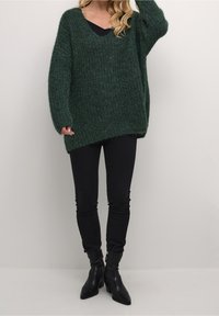 Grön stickad oversized tröja med V-ringning, kombinerad med svarta smala jeans och svarta spetsiga ankelboots. Mjuk textur och avslappnad passform.