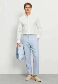 Weißes Langarmshirt mit Stehkragen, hellblaue maßgeschneiderte Hose und beige Slipper, hält einen hellblauen Blazer in der Hand.