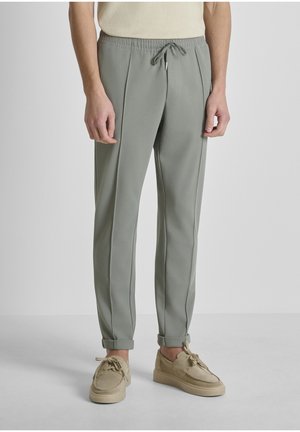 Uomo indossa pantaloni jogger affusolati grigio chiaro con coulisse e scarpe casual beige con lacci, in piedi su uno sfondo neutro.