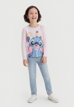 Jeune fille souriante, portant un t-shirt à manches longues rose clair avec le personnage de dessin animé Stitch en bleu, et un jean bleu clair, debout devant un fond blanc.