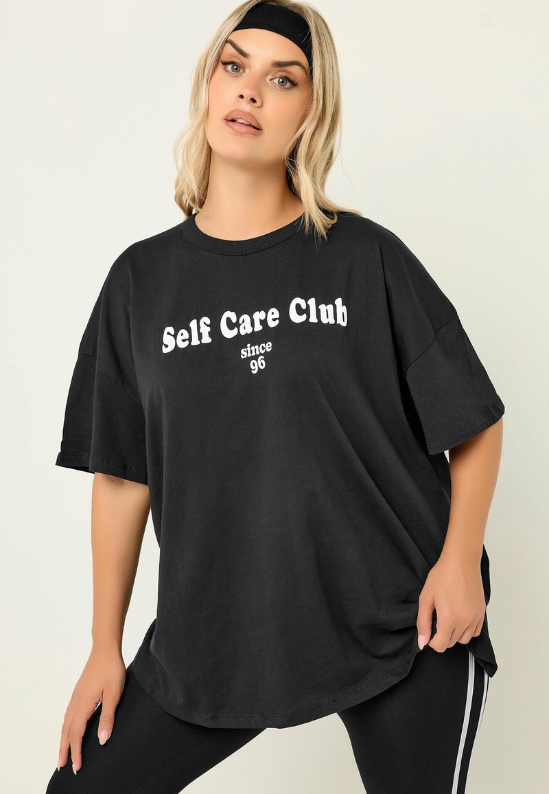 Frau trägt ein schwarzes, übergroßes T-Shirt mit dem Text "Self Care Club seit 96", schwarzes Stirnband und schwarze Leggings mit weißen Streifen.