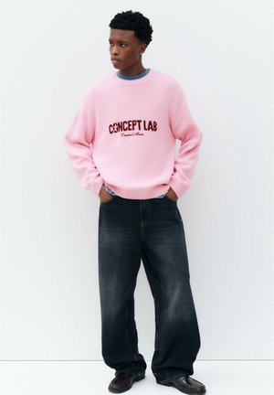 Ung mand stående mod en hvid væg iført en lyserød "CONCEPT LAB" sweater, mørke vide jeans og sorte sko med hænderne i lommerne.