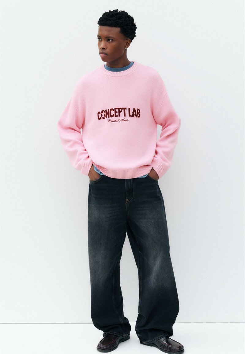 Jeune homme debout contre un mur blanc, portant un pull rose "CONCEPT LAB", un jean large foncé et des chaussures noires, les mains dans les poches.