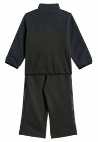 Černý sportovní oblek se zipem na fleecové bundě a odpovídajícími kalhotami. Fleecová textura na ramenou, jednobarevné černé kalhoty s vzorovanými bočními detaily.