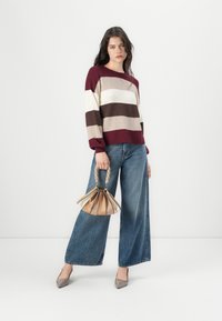 Pull en tricot rayé bordeaux, beige et marron ; jean denim à jambes larges ; sac à main beige avec une poignée en chaîne ; talons taupe.