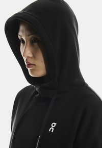 Mustar-kaputõmblus, millel on paelad ja rindkere peal väike valge logo. Pehme tekstuur ja näo ümber sobiv disain.