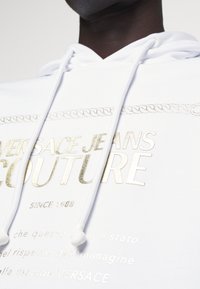Vit hoodie med "VERSACE JEANS COUTURE" i guldmetalliskt tryck. Inkluderar dragsko med vita spetsar och dekorativ kedjedesign.
