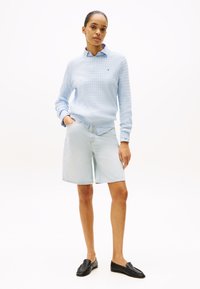 Lyseblå teksturert strikket genser med ribbestrikkede mansjetter, kombinert med lyse denimshorts. Modellen har på seg svarte loafers og står foran en hvit bakgrunn.