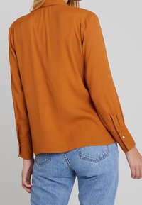 Rustorange skjorte med lange ærmer, krave og knapdetaljer ved manchetten. Båret med lyseblå denimjeans. Glat tekstur.