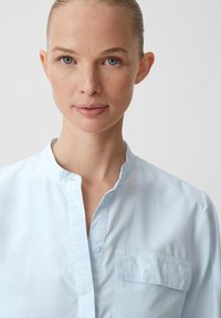 Jasnoniebieska bawełniana koszula z kołnierzykiem typu band i zapięciem na guziki, wyposażona w kieszeń na piersi i o miękkiej fakturze.