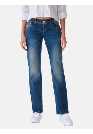 Femme portant un jean droit bleu délavé, des baskets blanches et une chemise blanche courte à boutons avec les manches retroussées.