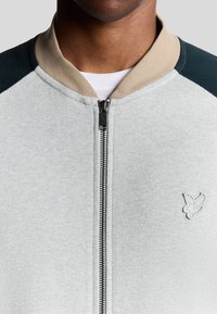 Helles graues Zip-Up-Sweatshirt mit beigen und dunkelgrünen Einsätzen, einem Stehkragen und einem gestickten Logo auf der linken Brust. Weicher Textil.
