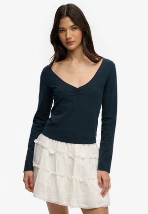 Mujer con cabello largo y oscuro, vestida con una blusa azul marino de manga larga y cuello en V, y una falda blanca de varios volantes, posando contra un fondo blanco liso.