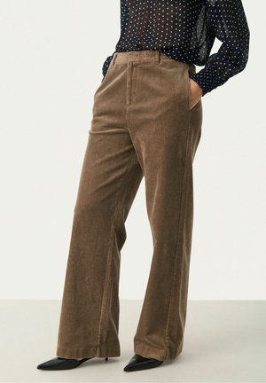 Pantalon large en velours côtelé marron porté avec des talons noirs à bout pointu et un chemisier bleu marine transparent à pois, les mains dans les poches.