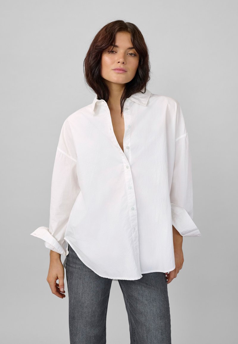 White button-up shirt with a loose fit, rolled sleeves, and light blue buttons. Paired with dark gray jeans. Smooth, lightweight fabric.