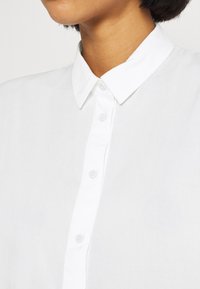 Camicia bianca con bottoni, in tessuto liscio, colletto classico e bottoni piccoli, che presenta un design pulito e semplice.