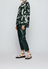 Blouse à manches longues, verte à motifs floraux avec des motifs abstraits blancs, associée à un pantalon en cuir vert ajusté et des sandales blanches avec une semelle noire.