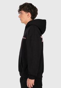 Young man in black hoodie standing sideways against plain light background, with hood up and pink text partially visible on chest and sleeve.