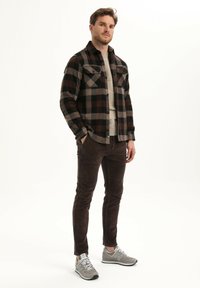 Flanellen overhemd in zwart en bruin ruitpatroon, beige trui, bruine corduroy broeken en grijze sneakers. Zichtbare knoopdetails en zakken.