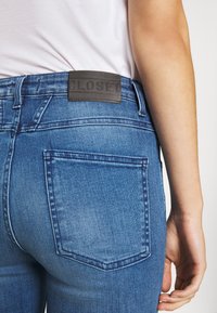Jean denim bleu avec une finition délavée, comprenant un patch en cuir étiqueté « CLOSED », des poches arrière visibles, et des détails de couture standard.