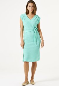 Mintgroene jurk tot de knie met een V-hals, kapmouwen en een gestrikte tailleband. Getextureerde stof met een zijsplitdetail.