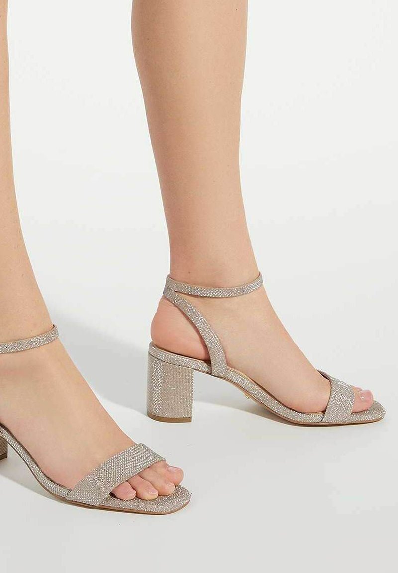 Glanzende zilveren sandalen met hakken hebben een stevige blokhak, open teen en enkelbandje. De banden hebben een gestructureerde afwerking voor extra detail.