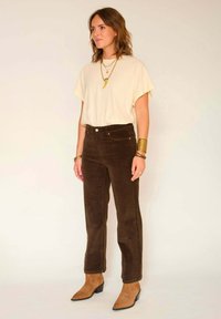 Chemise crème à manches courtes, pantalon en velours côtelé marron taille haute, et bottines beiges à bouts pointus. Accents de bijoux en or visibles.