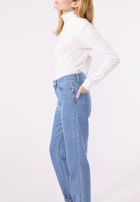 Frau, die einen weißen Rollkragenpullover und hochgeschnittene blaue Jeans trägt, mit einer Hand in ihrer Tasche, vor einem weißen Hintergrund stehend.
